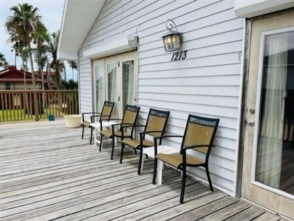 A wooden deck with four chairs and a lantern, number 1213 on the wall, palm trees in the background.