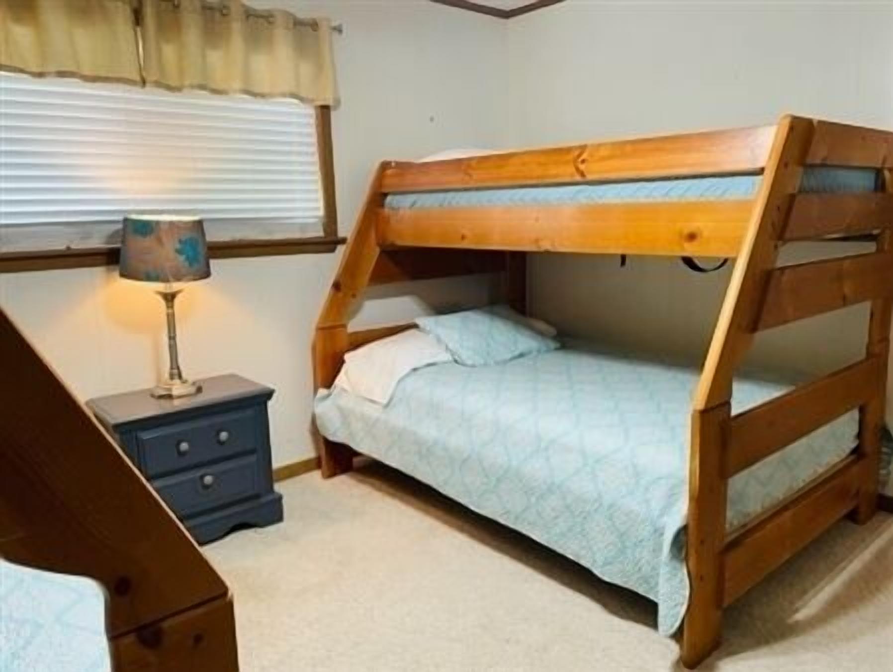 A bunk bed with a wooden frame and a blue mattress, a bedside table with a lamp, and a window with blinds.