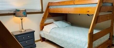 A bunk bed with a wooden frame and a blue mattress, a bedside table with a lamp, and a window with blinds.