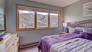 Une chambre avec une grande fenêtre, un lit avec une literie violette, une table de chevet avec une lampe et une commode.