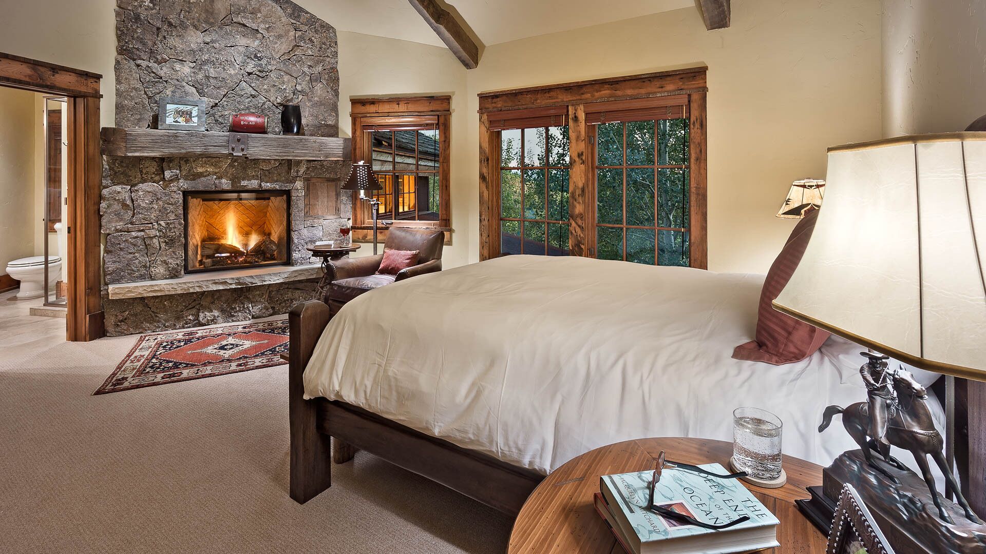 A bedroom with a stone fireplace, a large bed, a wooden table with a book and glasses, and a view of trees through a window.
