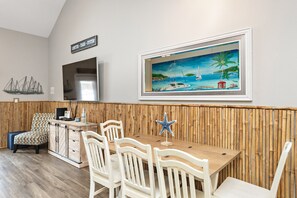 A dining area with a wooden table and white chairs, a framed painting of a beach scene, and a bamboo wall panel. - Poolside Paradise | Private Patio & Pet-Friendly (Corpus Christi)