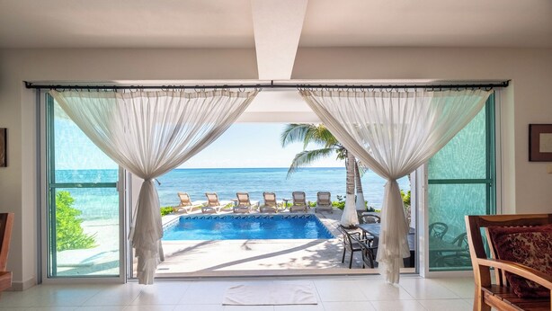 A room with a large sliding glass door leading to a pool area with lounge chairs and a view of the ocean. - Spacious Beachfront Living on Akumal Bay (South Akumal)