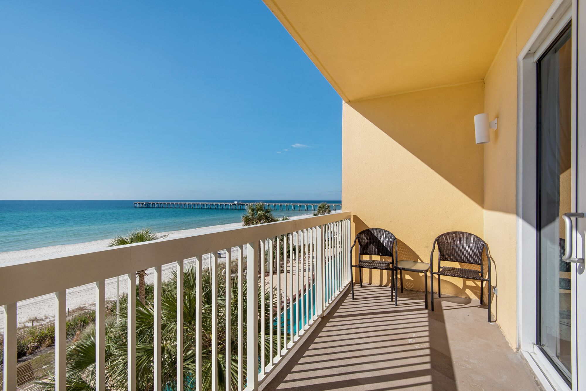 A balcony with a view of the ocean and a pier, featuring two wicker chairs and a white railing.
