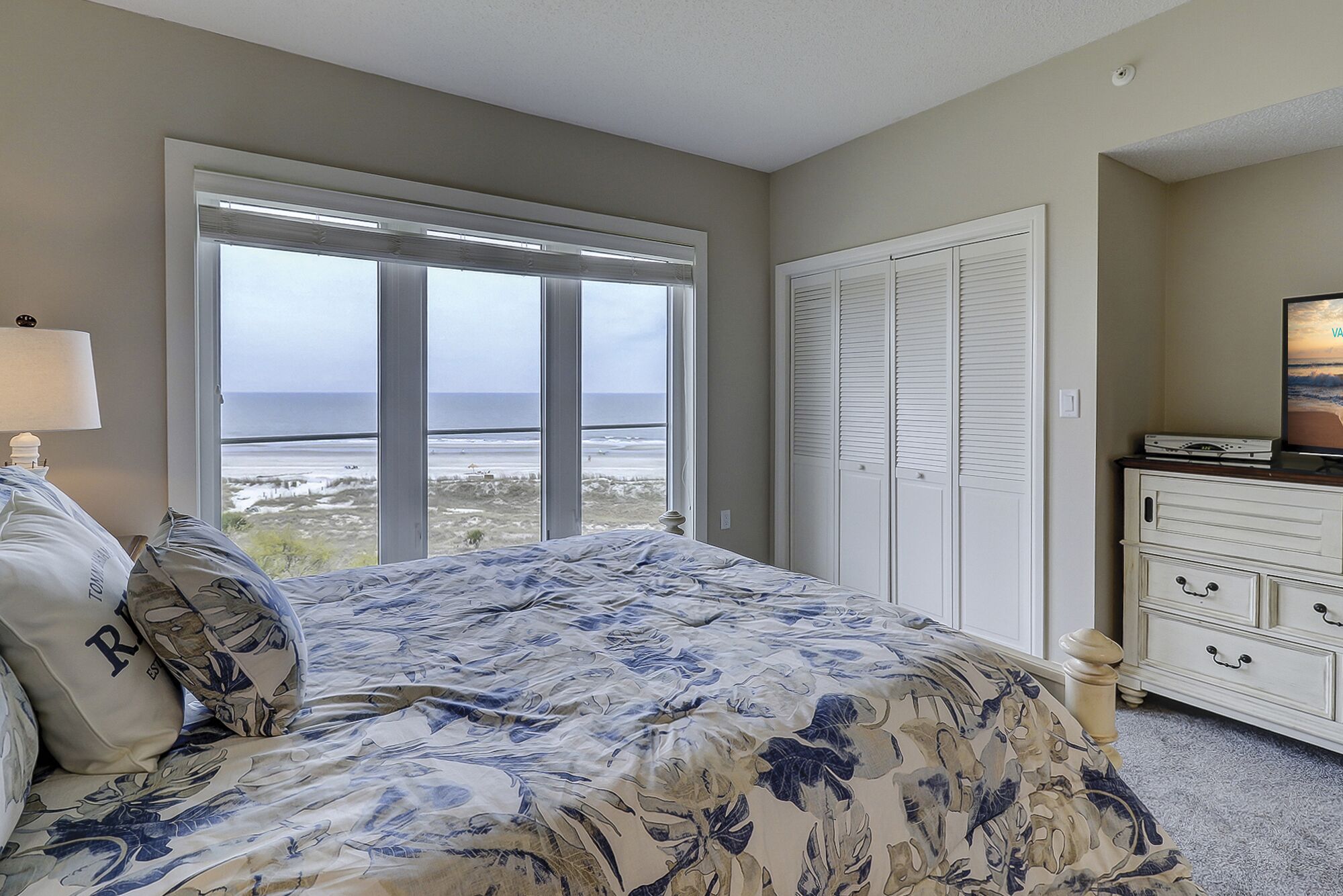 Een slaapkamer met een groot raam dat uitkijkt op het strand, een bed met bloemenbedrukking, een dressoir en een televisie op het dressoir.