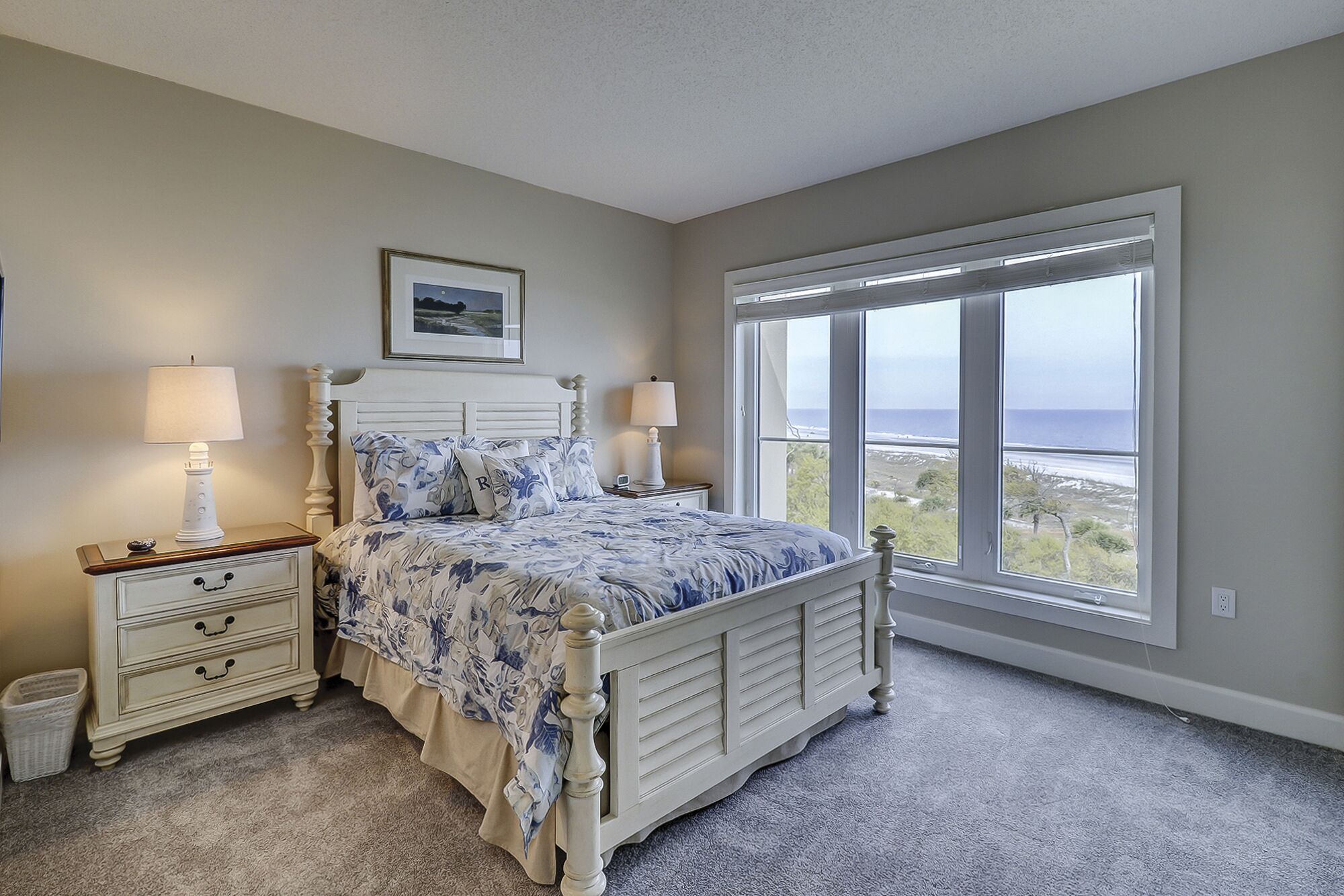 A bedroom with a bed, two bedside tables with lamps, a framed picture on the wall, and a large window with a view of the ocean.