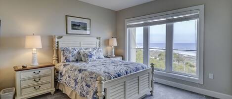 A bedroom with a bed, two bedside tables with lamps, a framed picture on the wall, and a large window with a view of the ocean.