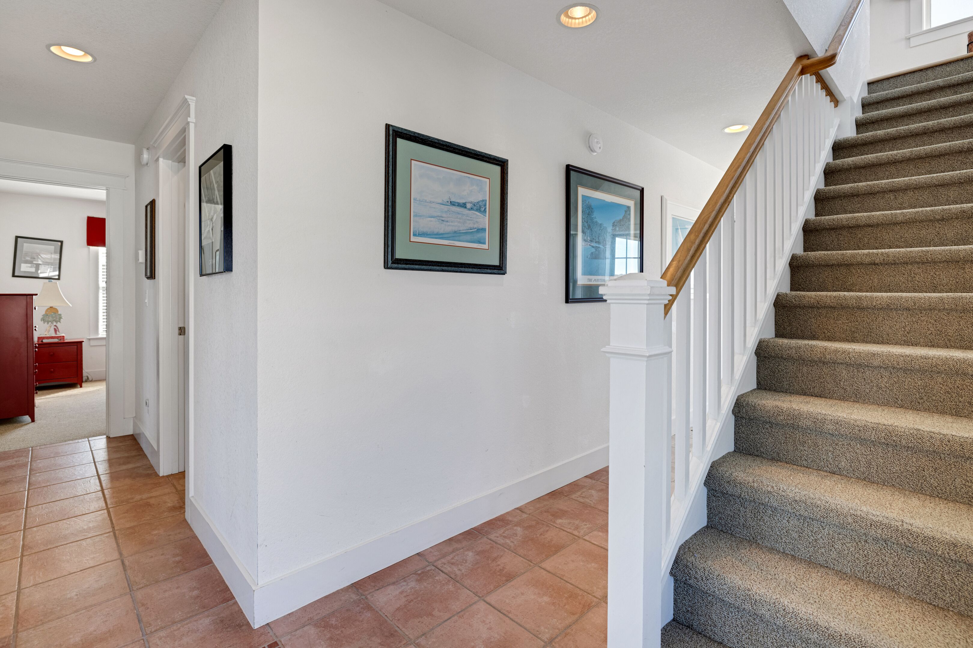 A hallway with a staircase, white walls, and framed pictures.