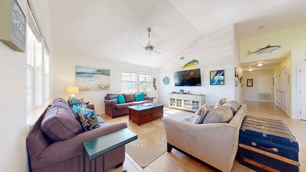 A spacious living room with a high ceiling, a ceiling fan, and a large TV mounted on the wall. There is a sofa, a coffee table, and a rug on the floor. - Parking for Boat, Shared Pool, Outdoor Seating Areas, Pet Friendly (Port Aransas)