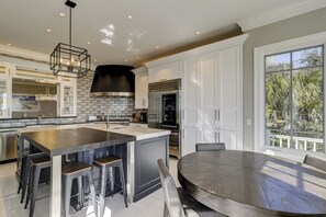 A modern kitchen with a central island, bar stools, a round dining table, and a large window with a view of greenery. - 33 Dune Lane- Oceanfront & Beautiful! (Hilton Head Island)