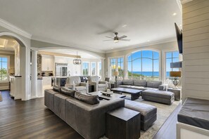 A spacious living area with a large sofa, a coffee table, and a television. There is a kitchen in the background with white cabinets and a view of the ocean. - 33 Dune Lane- Oceanfront & Beautiful! (Hilton Head Island)