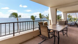 A balcony with a table and chairs, overlooking the ocean with palm trees and a clear sky.