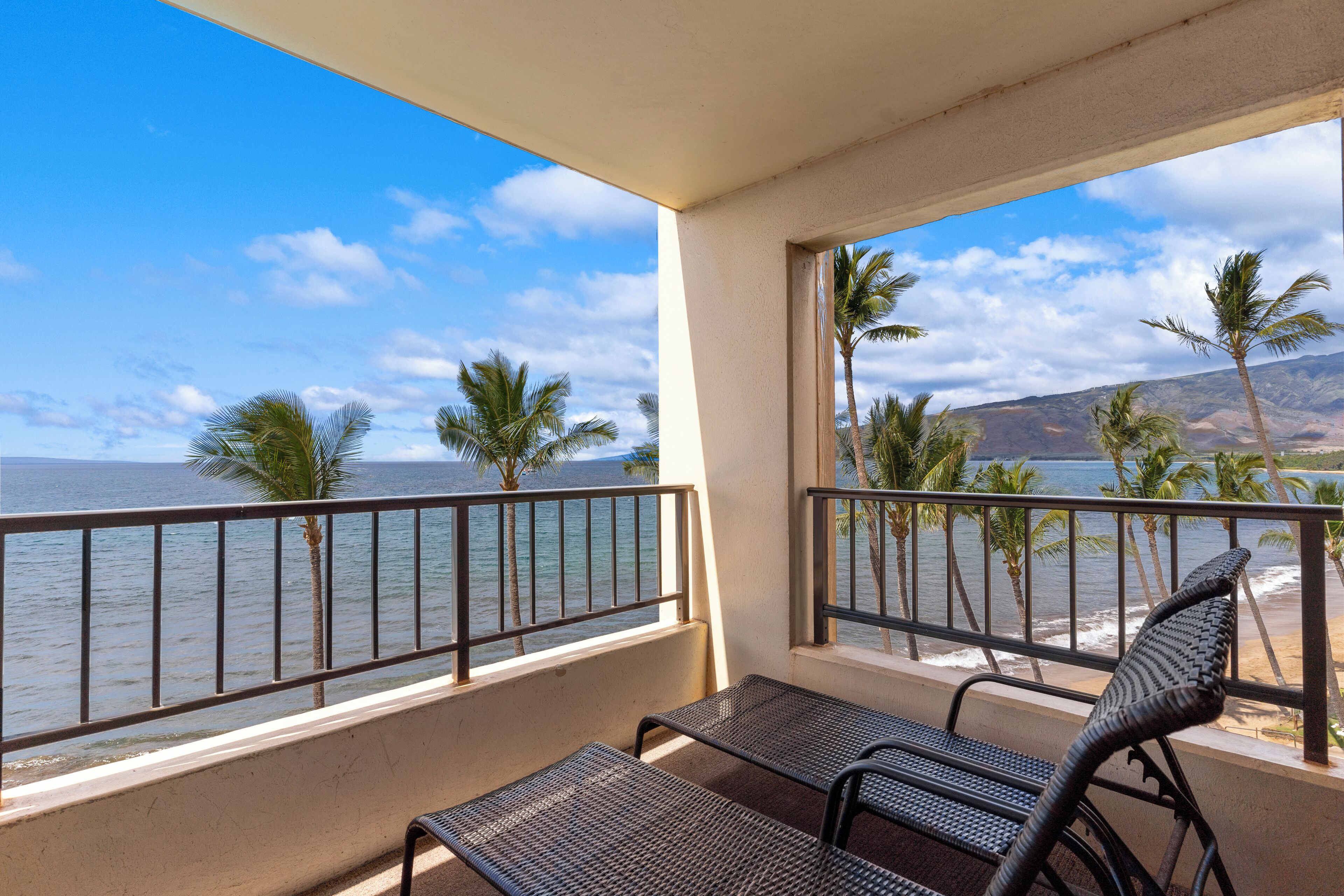 A balcony with two wicker chairs, a view of the ocean, and palm trees.