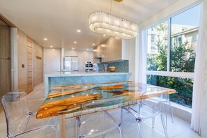 A modern kitchen with a glass-top dining table, clear chairs, and a large window with a view of greenery.