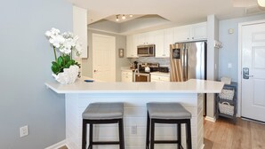 A kitchen with white cabinets, a stainless steel refrigerator, a microwave, and a sink. There are two bar stools in front of the counter. - Fully equipped, colorful, bright condo w/private balcony! (Destin)