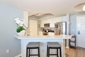 A kitchen with white cabinets, a stainless steel refrigerator, a microwave, and a sink. There are two bar stools in front of the counter.
