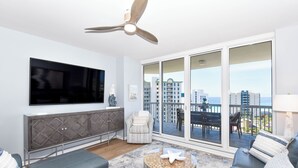 A modern living room with a flat-screen TV, a ceiling fan, a sofa, a coffee table with decorative items, and a balcony with a view of the city. - Fully equipped, colorful, bright condo w/private balcony! (Destin)