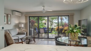 A modern living room with a sofa, coffee table, and chairs, a dining table with a vase of flowers, and a view of the outdoors through large sliding glass doors.