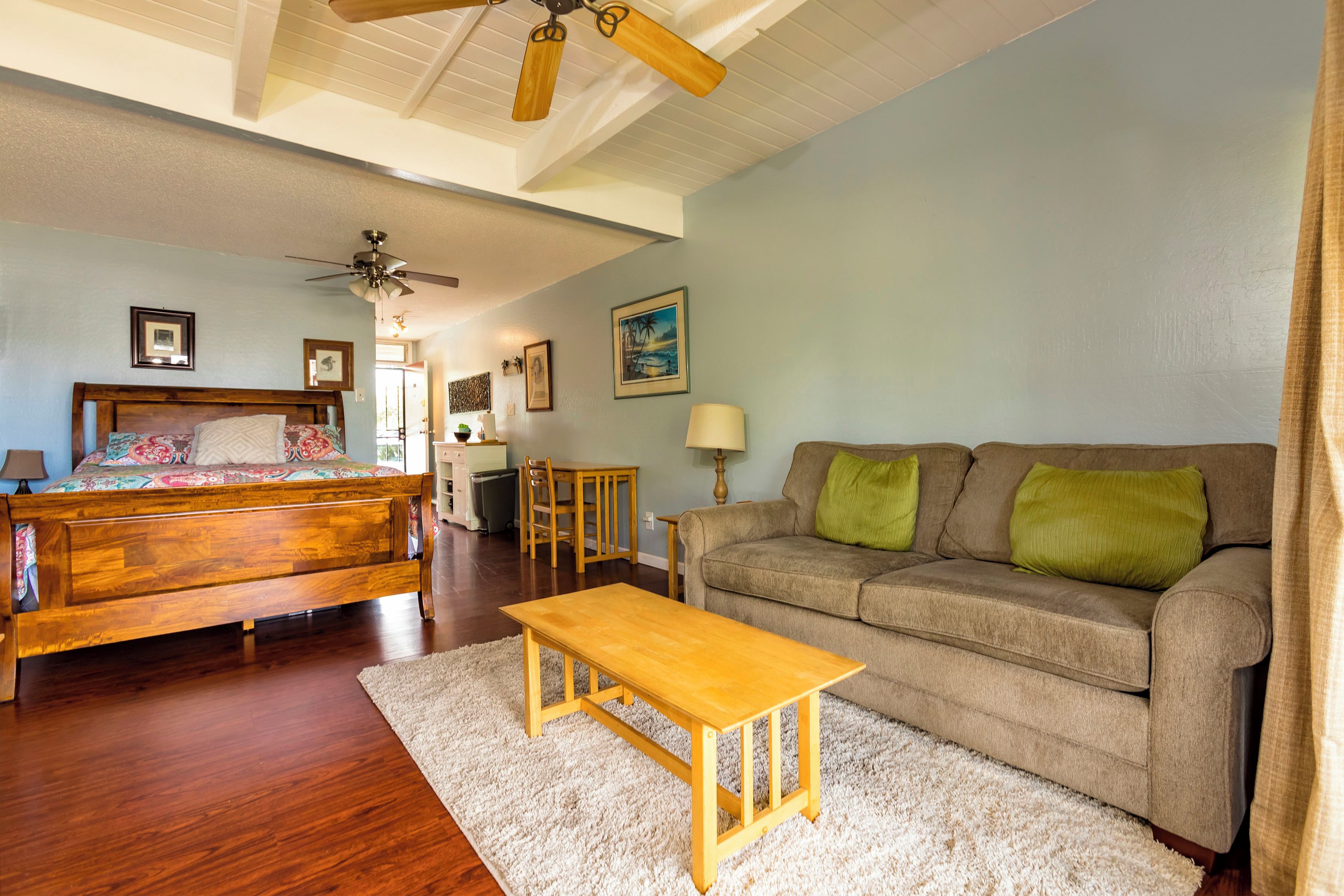 A spacious living room with a wooden bed, a wooden coffee table, a grey sofa with green cushions, and a ceiling fan.