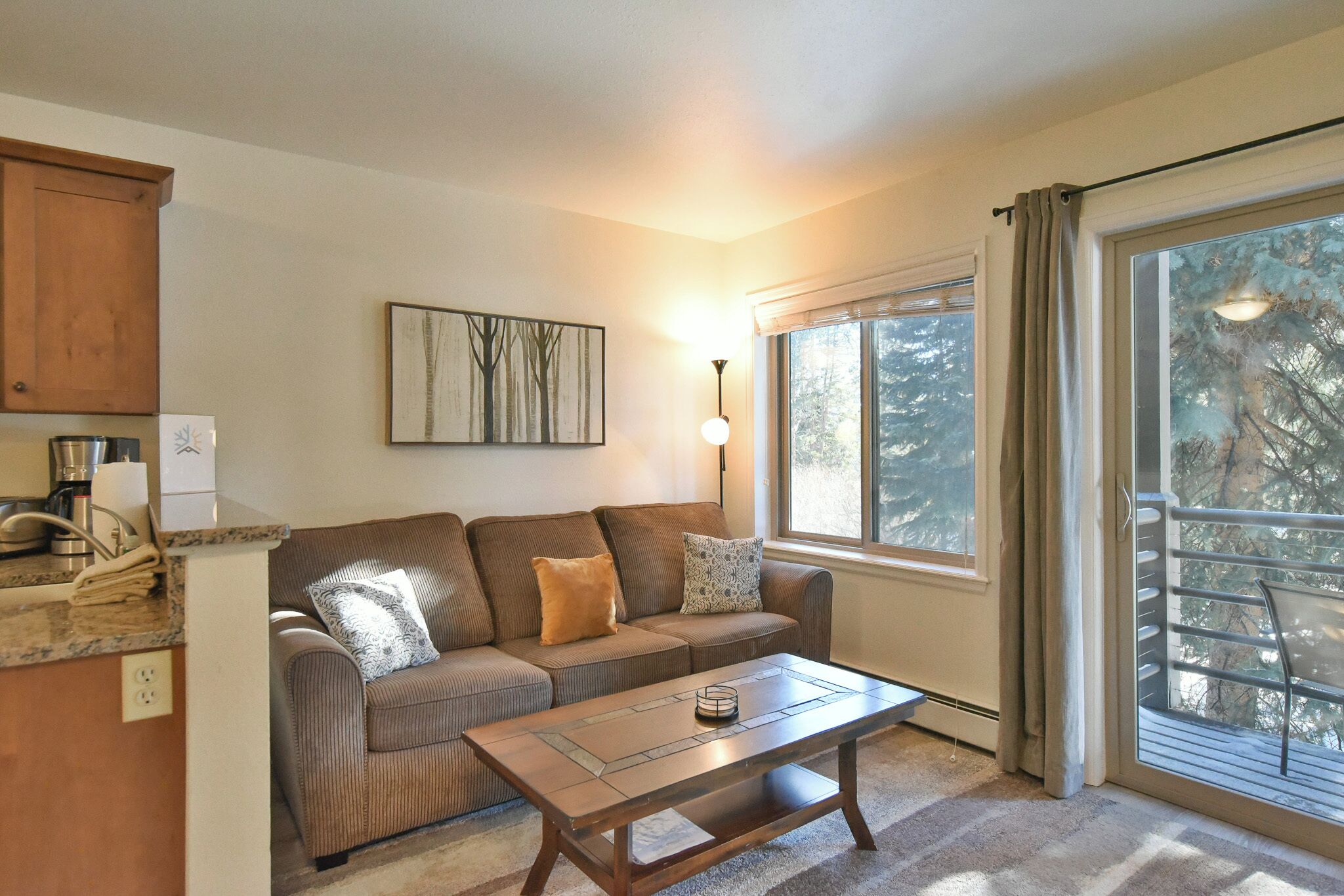 A cozy living room with a brown sofa, a coffee table, a painting on the wall, and a window with a view of trees.