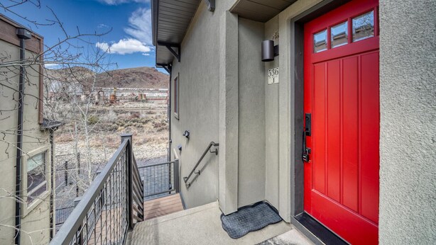 A red door with a mailbox and a number sign, a staircase leading to a balcony, and a view of a barren landscape with distant buildings.