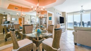 A spacious dining area with a glass-top table surrounded by wicker chairs, a chandelier, and a view of the ocean through large windows. - Coastal Beachfront Condo w/ Relaxing Community Pool in Destin! 💙☀🌊 (Destin)