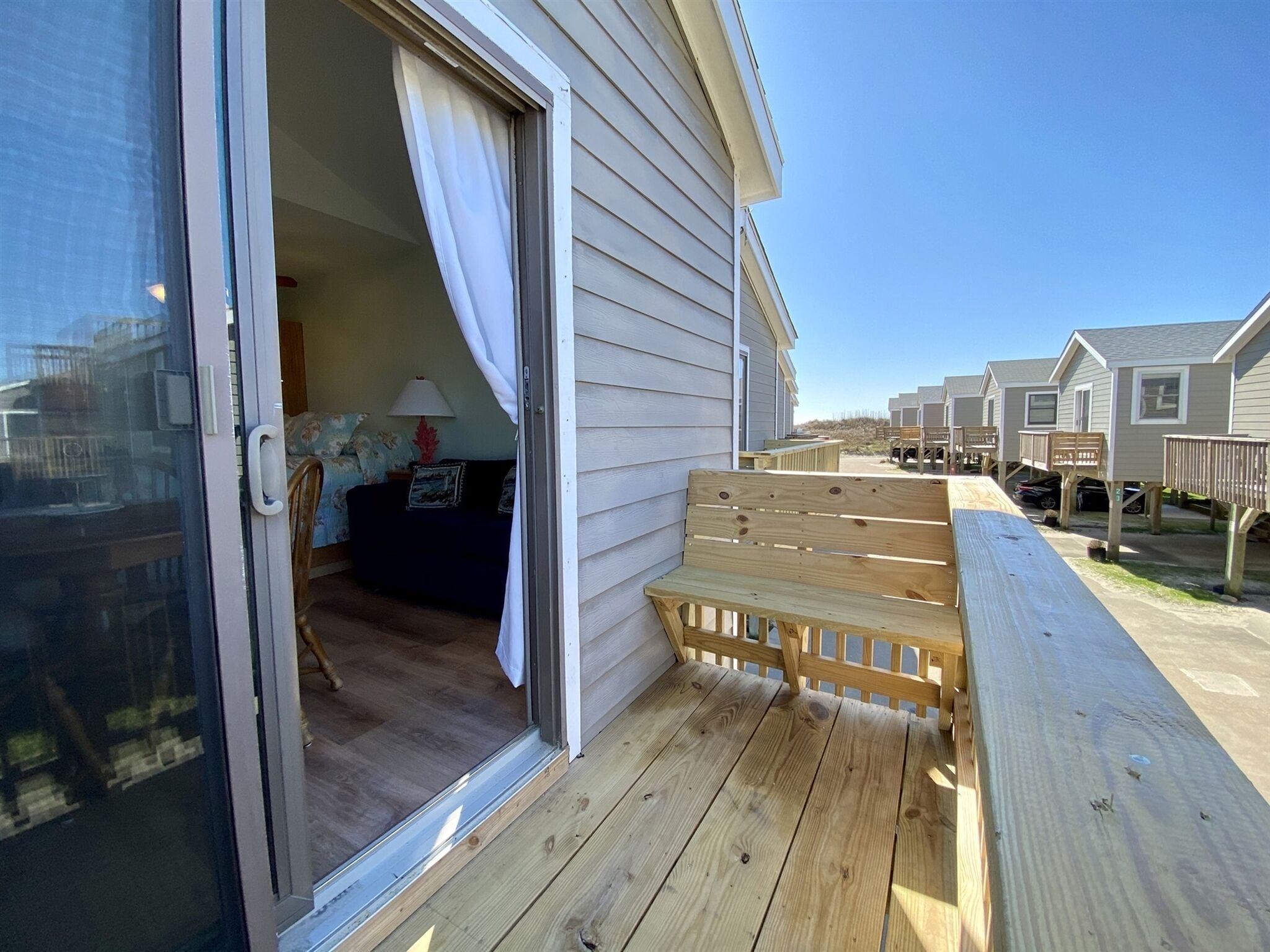 A wooden deck with a bench, a sliding glass door leading to a bedroom with a bed and a nightstand, and a view of other similar buildings in the background.