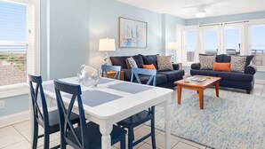 A living room with a white table and blue chairs, a dark blue sofa with patterned cushions, and a painting on the wall.
