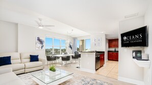 A modern living room with a sofa, coffee table, and a dining area with chairs. There is a kitchen with a TV displaying "CEANS LUXURY VACATIONS" and a sign.