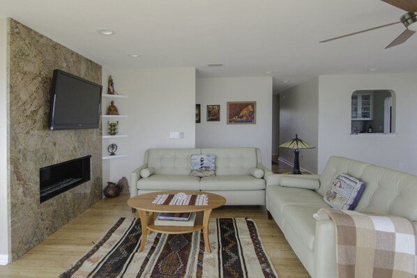 A living room with a sofa, a coffee table, a TV mounted on a stone wall, and a ceiling fan. - Large, newly renovated oceanfront home with superb views of Breach Inlet (Sullivan's Island)
