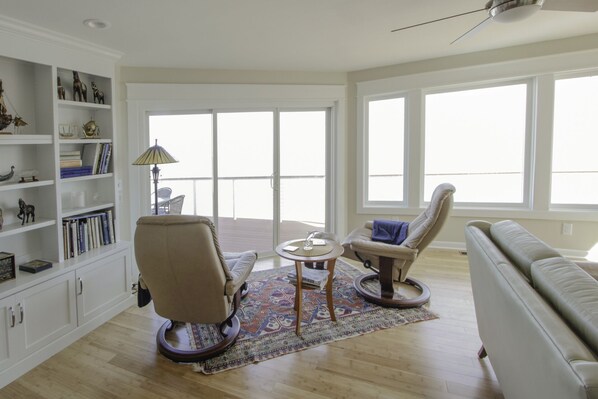 A spacious living room with a large window, a bookshelf, a sofa, and a chair. - Large, newly renovated oceanfront home with superb views of Breach Inlet (Sullivan's Island)