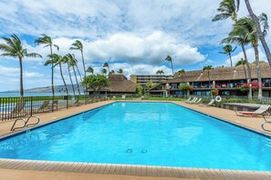 Ein rechteckiger Swimmingpool mit Holzterrasse, umgeben von Liegestühlen und Palmen, mit einem Gebäude im Hintergrund.