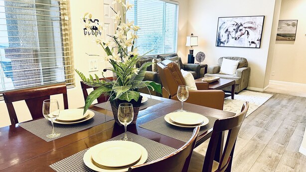 A dining area with a wooden table set for four, surrounded by chairs. There is a potted plant on the table, and a window with blinds to the left.