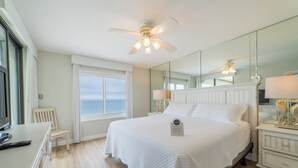 A bedroom with a large bed, a ceiling fan, a nightstand with a lamp, a chair, a desk with a remote control, and a window with a view.