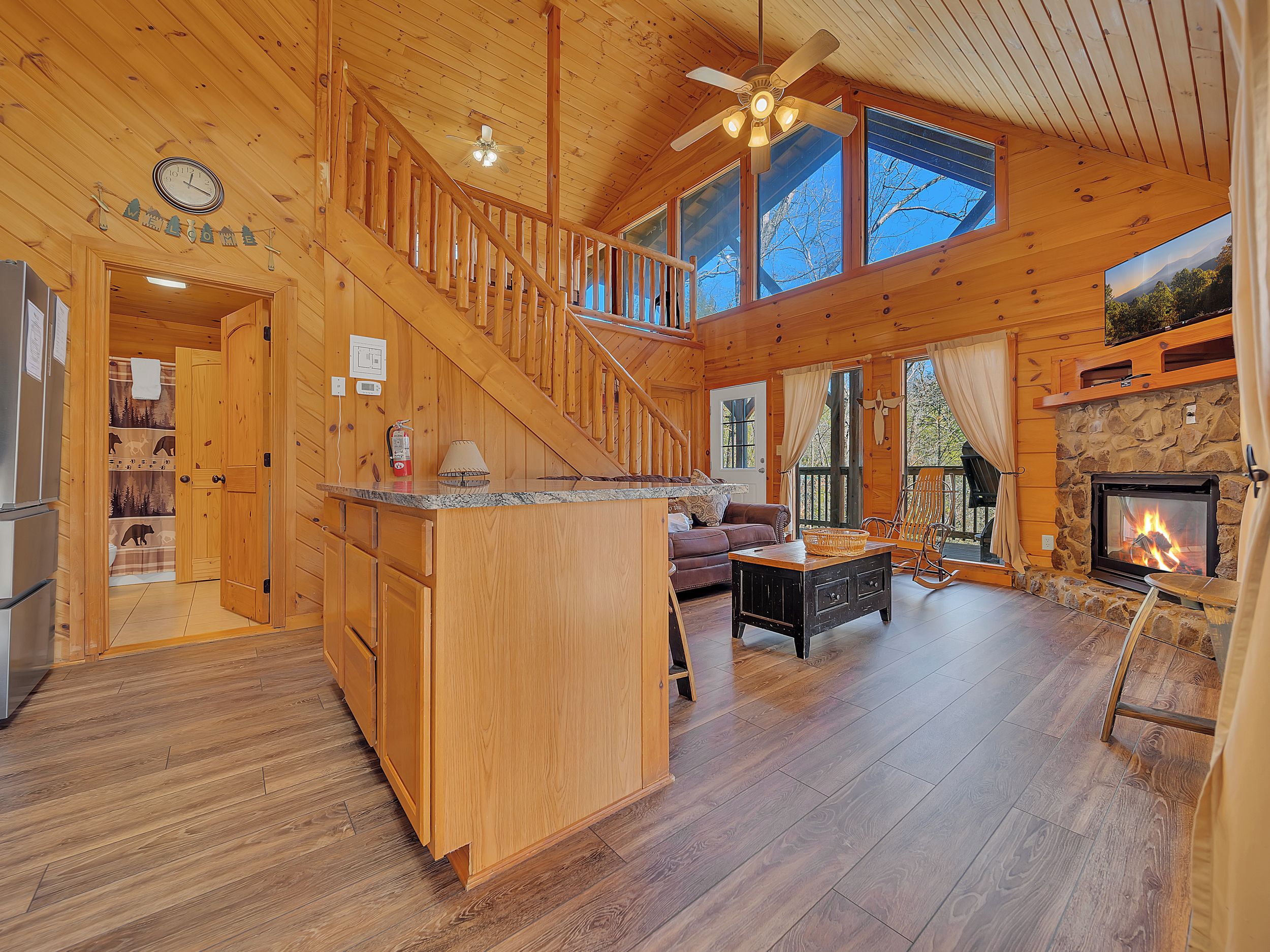 A wooden cabin interior with a central staircase, a fireplace, a dining area, and a kitchen.