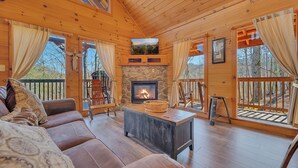 Interior acogedor de una cabaña con chimenea de piedra, piso de madera y un ventanal con vistas a una terraza.