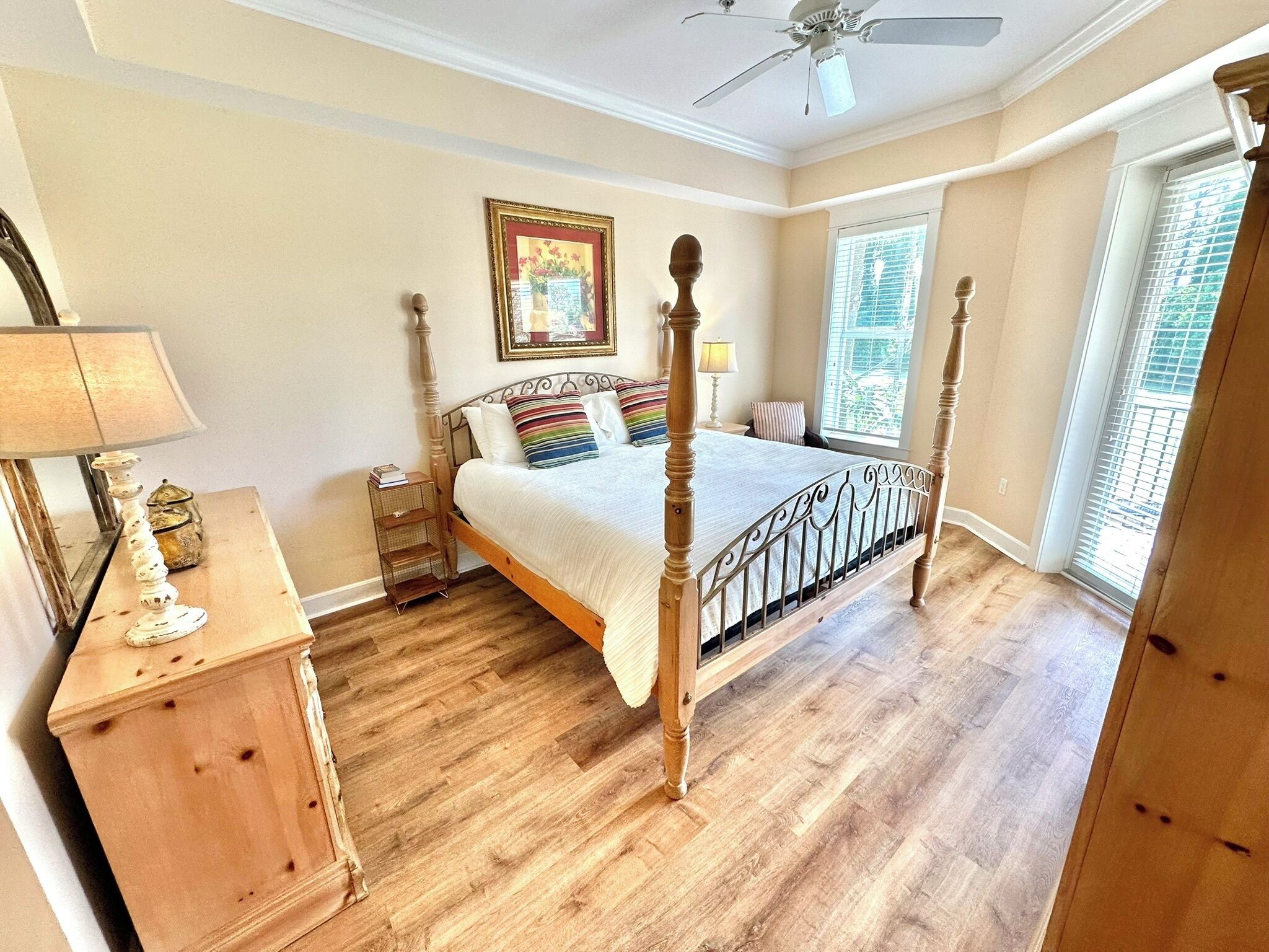 A bedroom with a four-poster bed, a nightstand, a lamp, a framed picture on the wall, and a window with blinds.