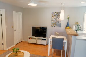 A modern living room with a flat-screen TV, a dining area with a table and chair, and a kitchen area with a sink and countertop.