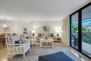 A spacious living room with a large window, a dining area with wooden furniture, and a balcony with a view of palm trees.