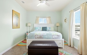 A bedroom with a bed, bedside lamps, a ceiling fan, a window with curtains, a colorful rug, and a wall-mounted picture.