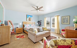 A bedroom with a bed, a sofa, a desk, a chair, a ceiling fan, and a window with curtains.