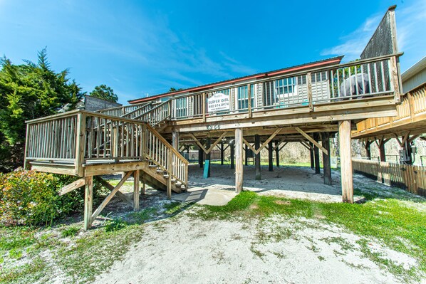 A wooden deck with a staircase leading to a covered area, surrounded by sand and sparse vegetation.