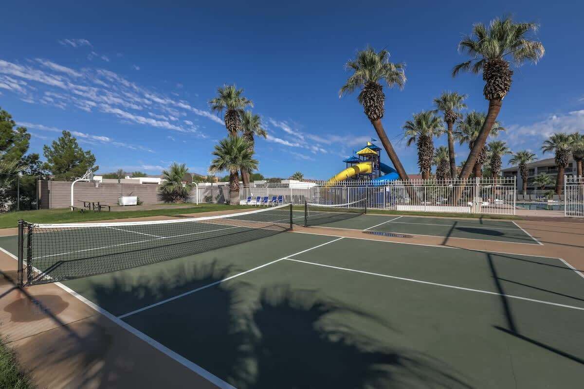 An outdoor tennis court with a net, surrounded by palm trees and a water slide in the background.