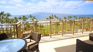 Un balcon doté de meubles en osier et d’une table en verre, offrant une vue sur une plage bordée de palmiers et sur l’océan.