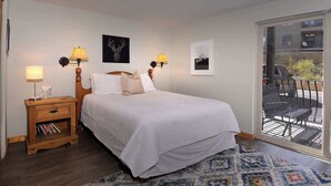 Una camera da letto con un letto, due lampade da comodino, un comodino con dei libri, un tappeto decorato e vista su un balcone esterno.