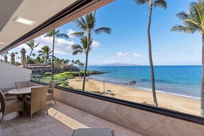 A balcony with wicker furniture overlooking a beach with palm trees and a clear blue sky.