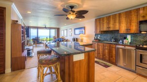 A modern kitchen with wooden cabinets, a central island with stools, stainless steel appliances, and a ceiling fan. - Aloha Condos, Keauhou Kona Surf and Racquet Club, Condo 1-102, Oceanfront (Kailua Kona)