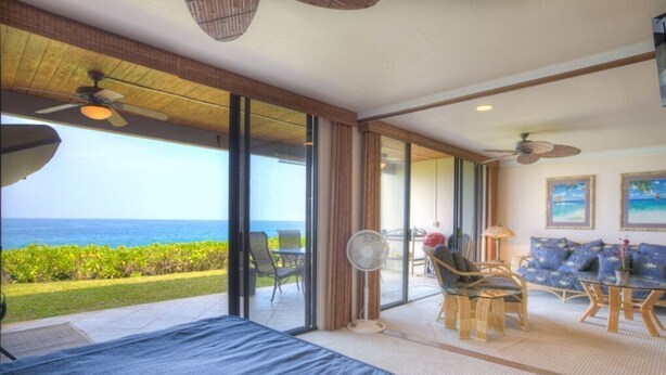 A room with a large sliding glass door leading to a balcony with a view of the ocean. There is a bed with a blue bedspread, a fan, and a table with chairs. - Aloha Condos, Keauhou Kona Surf and Racquet Club, Condo 1-102, Oceanfront (Kailua Kona)