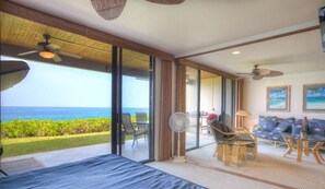 Une pièce dotée d’une grande porte-fenêtre coulissante donnant sur un balcon avec vue sur l’océan. On y trouve un lit avec une couvre-lit bleue, un ventilateur et une table avec des chaises.