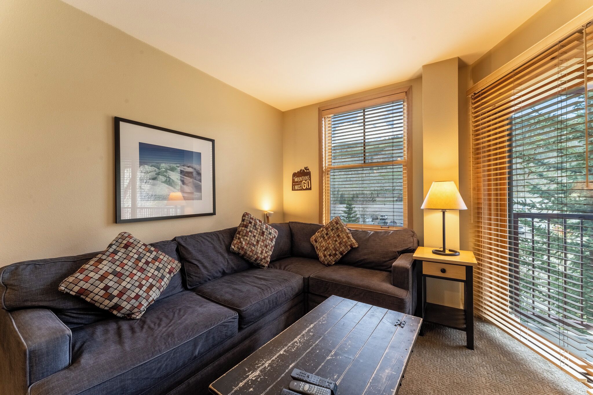 Sink into this cozy sectional with plaid pillows, where warm light filters through wooden blinds creating your perfect afternoon retreat.